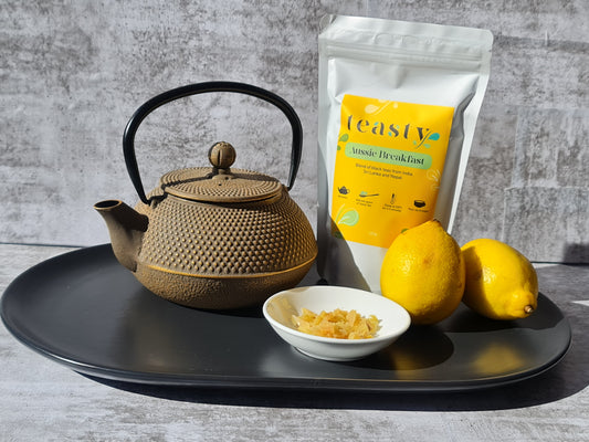 A cast iron tea pot sitting next to a packet of Teasty Aussie Breakfast Black Tea, a white dish of finely chopped ginger and two whole lemons. 